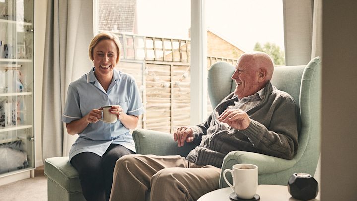 A carer and an older man share a laugh over cups of tea